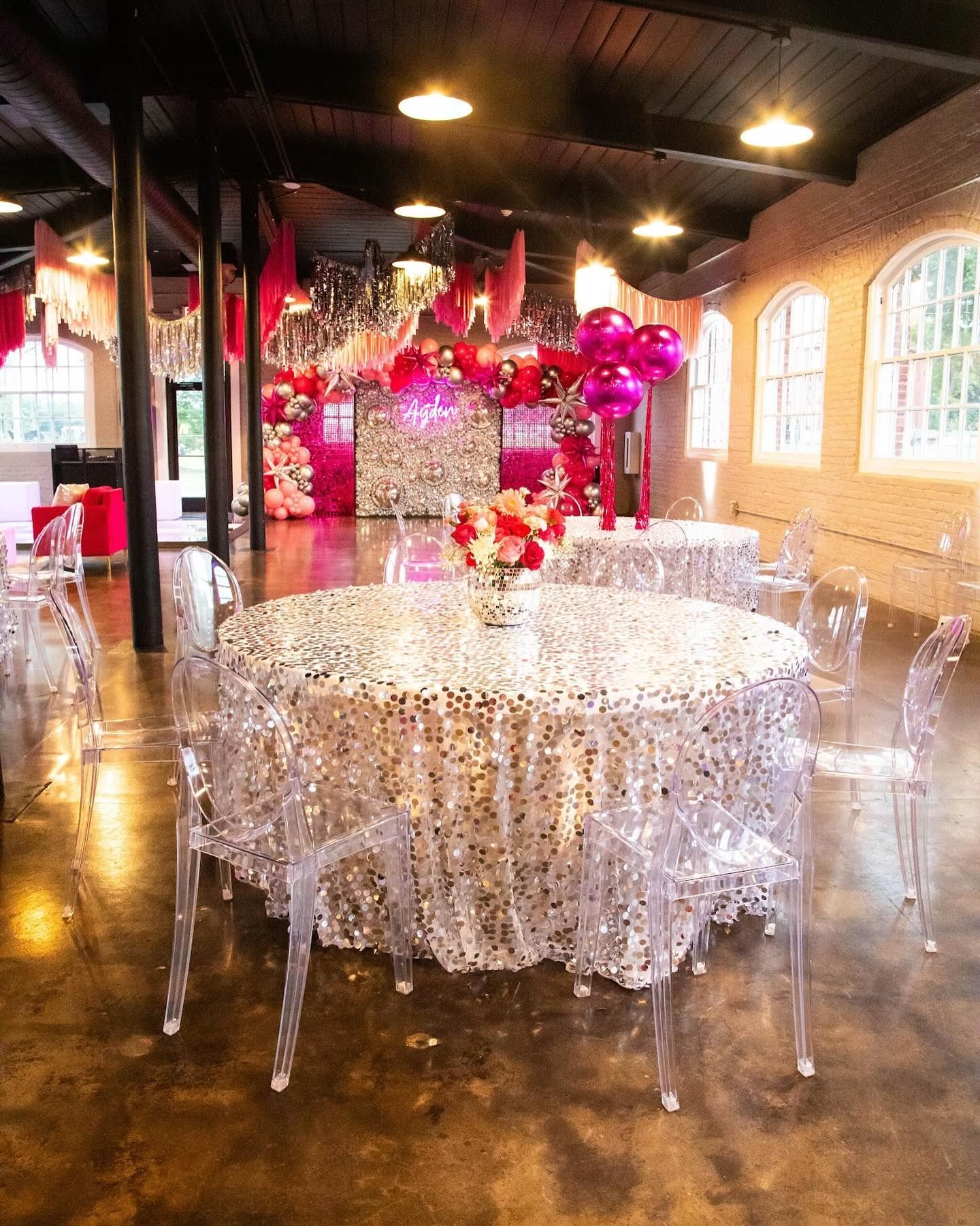 Indoor event space with round tables covered in sequined tablecloths and clear chairs, pink decor.
