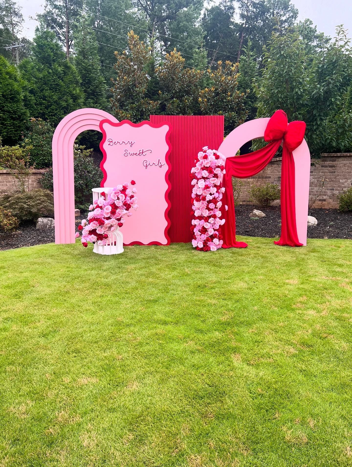 Pink and red party backdrop with flowers and archways set on green grass.