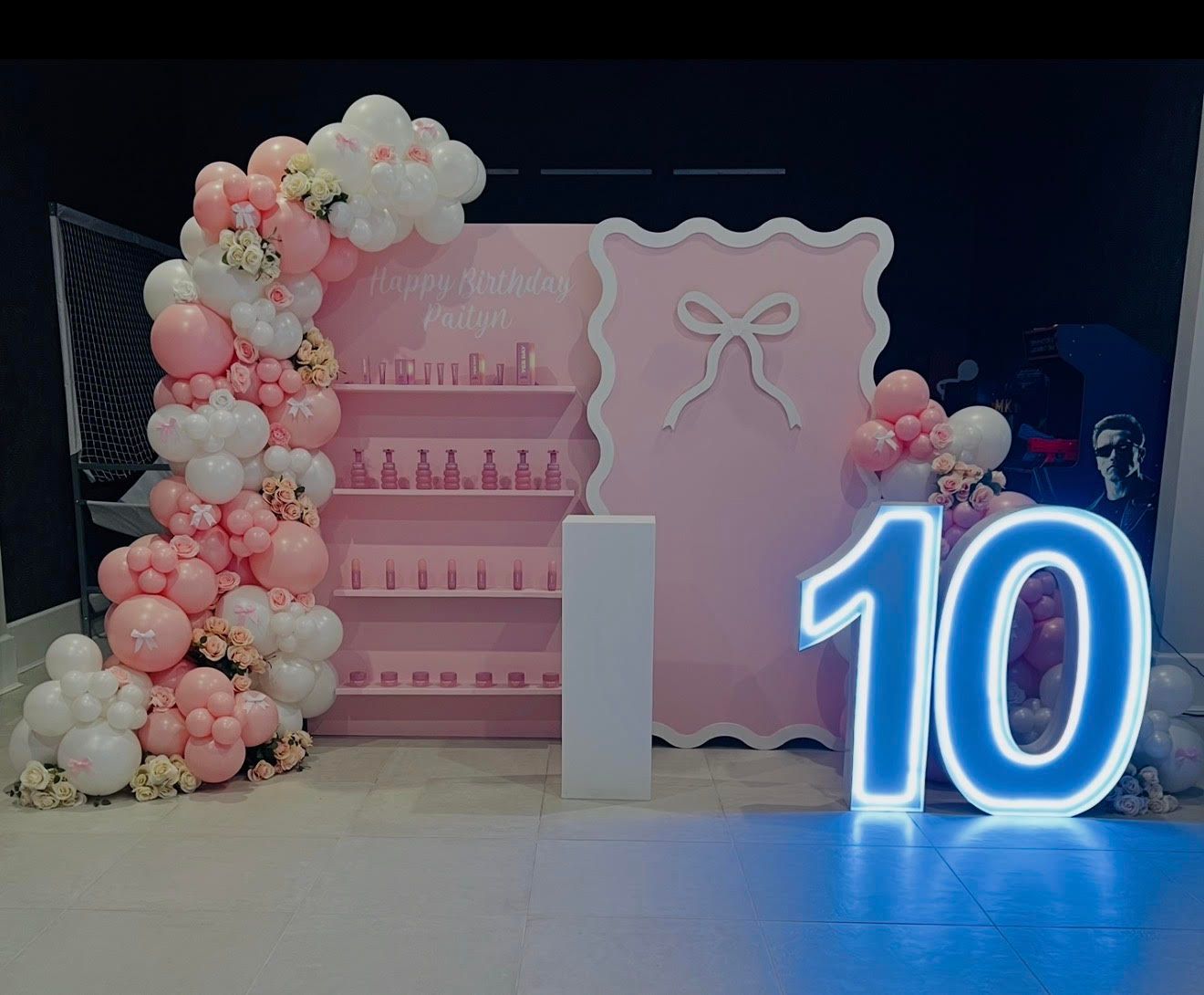 Pink and white balloon arch decorates a party backdrop with a pink wall, shelf display, and a neon