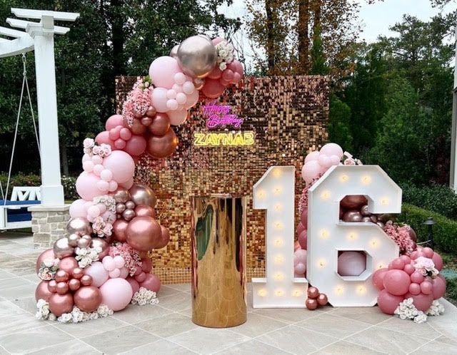 Rose gold 16th birthday party setup with balloon arch, sequin backdrop, and illuminated numbers.