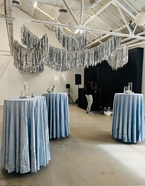 Three round tables with light blue cloths in a decorated event space with a DJ. Silver streamers hang from the ceiling.