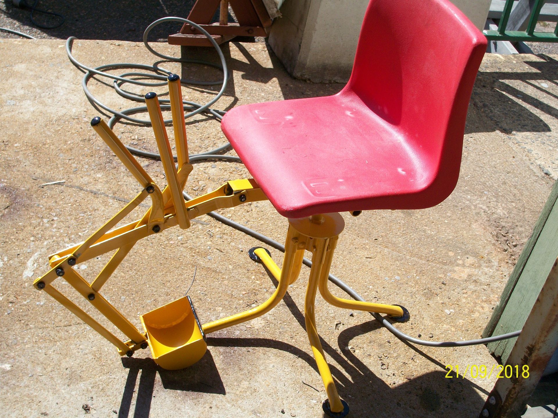Red chair mounted on a yellow metal pedal base outdoors on a concrete patio.