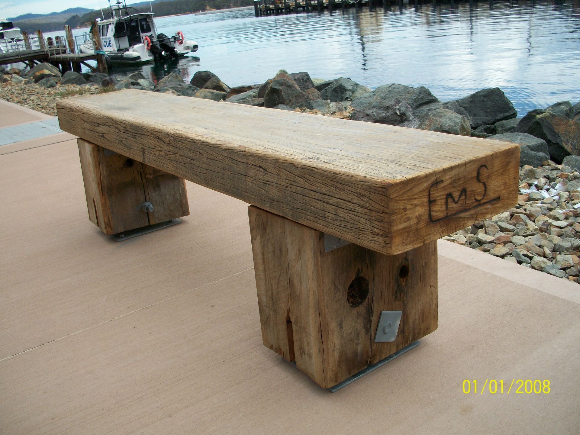Wooden bench on a waterfront promenade beside rocks and calm water, with boats in the background.