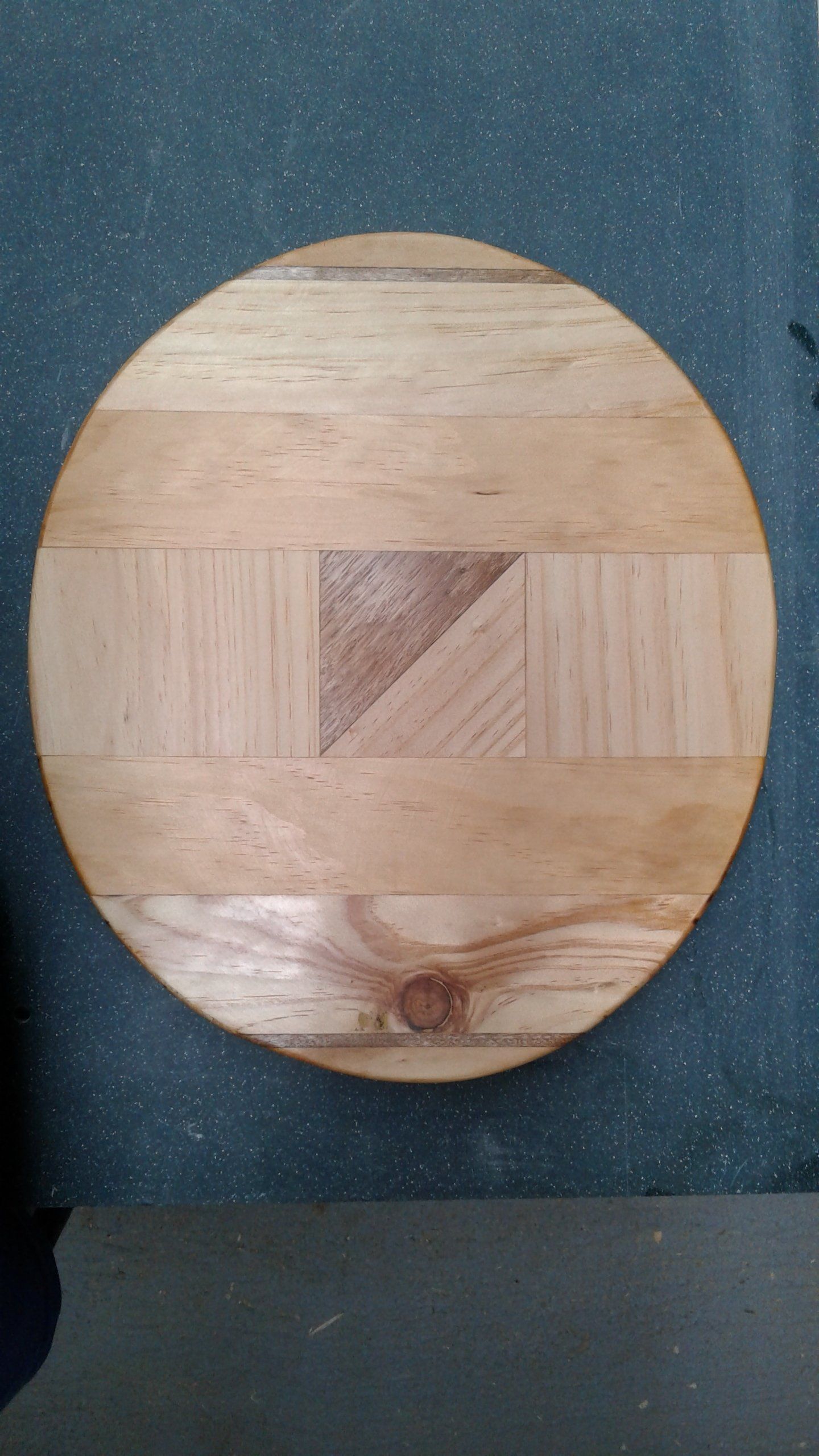 Round wooden cutting board with mixed light and dark wood grain, centered on a dark speckled surface