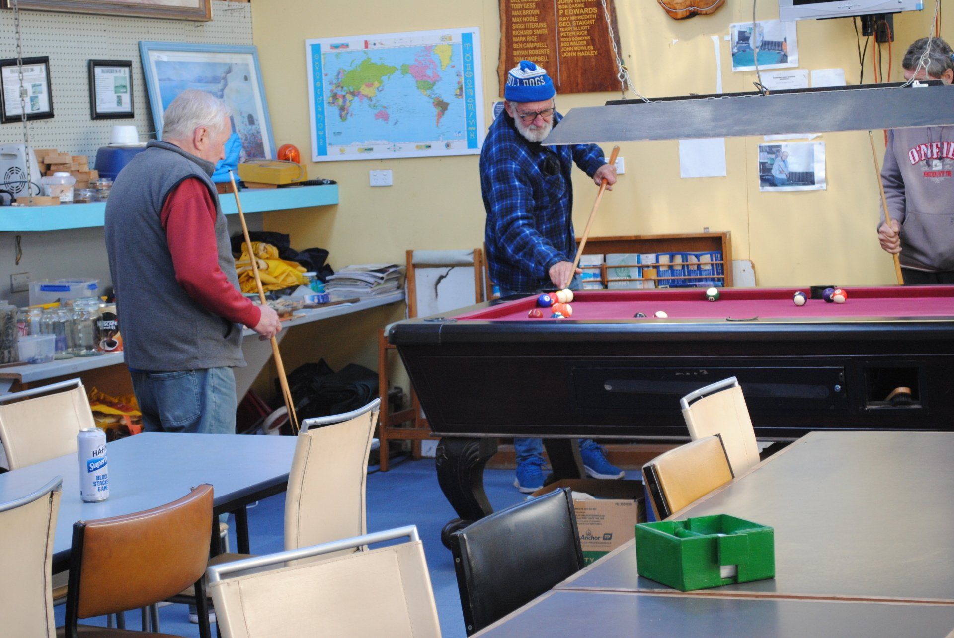 People playing pool in a recreation room with tables, chairs, and wall maps.