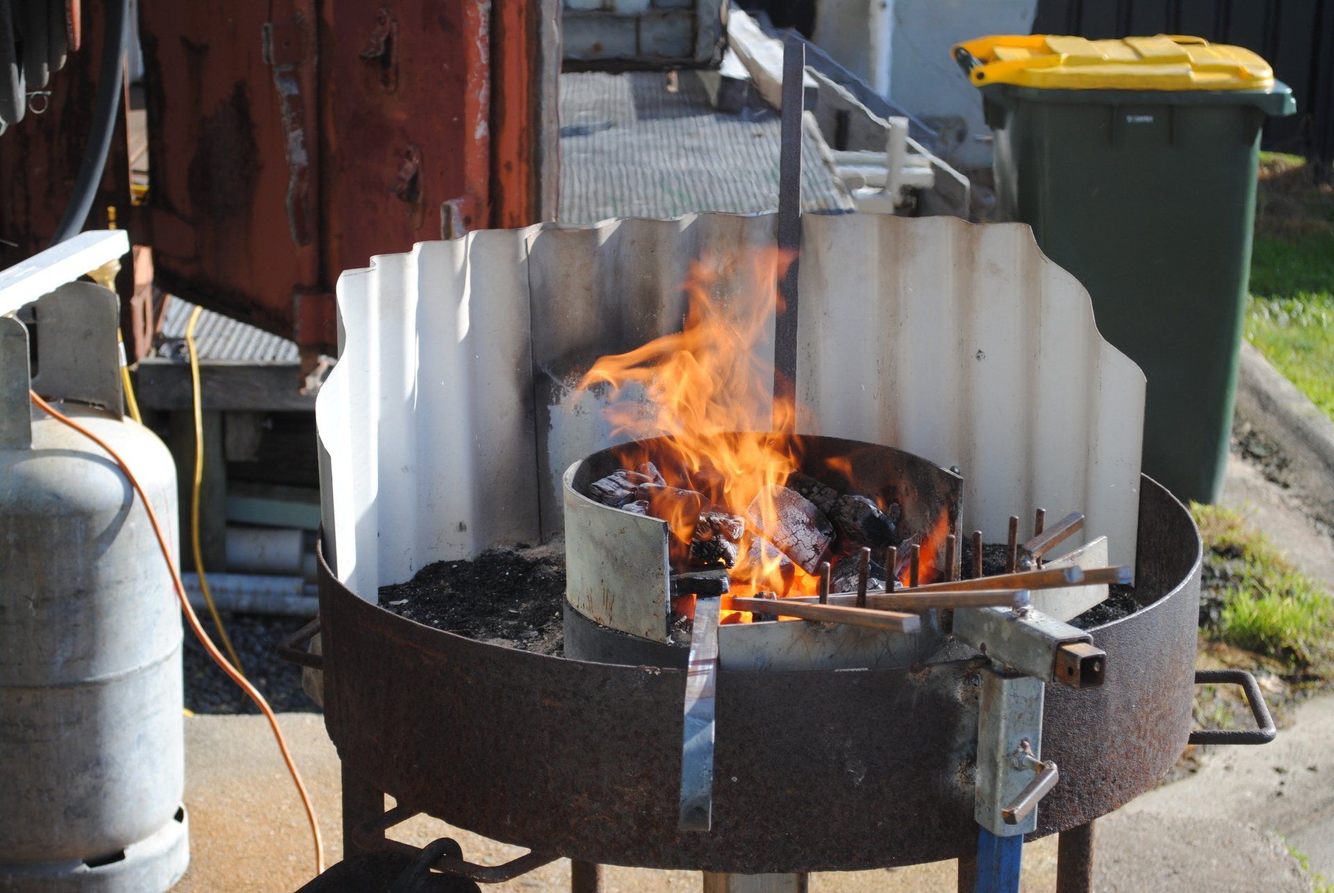 Small outdoor fire burning in a round metal pit with corrugated windbreak panels and charcoal