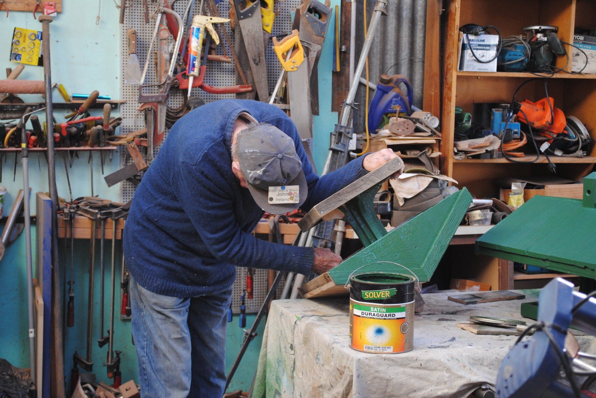 Person sanding a green panel in a cluttered workshop with tools and paint cans nearby