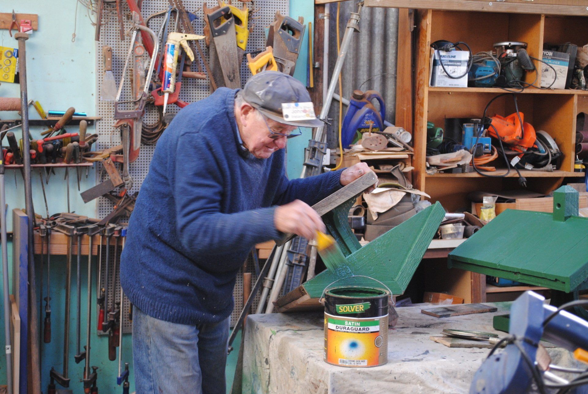 Man working in a cluttered workshop, painting a green object on a bench.