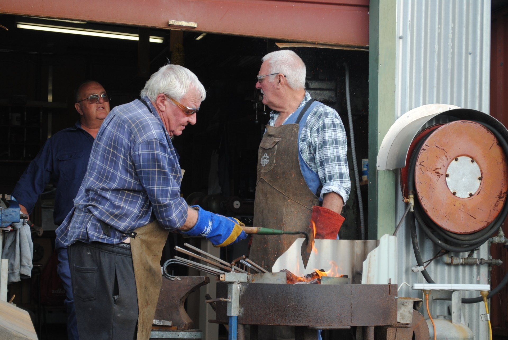 Two men in work gloves tending a metal furnace in a workshop, with orange flames visible.