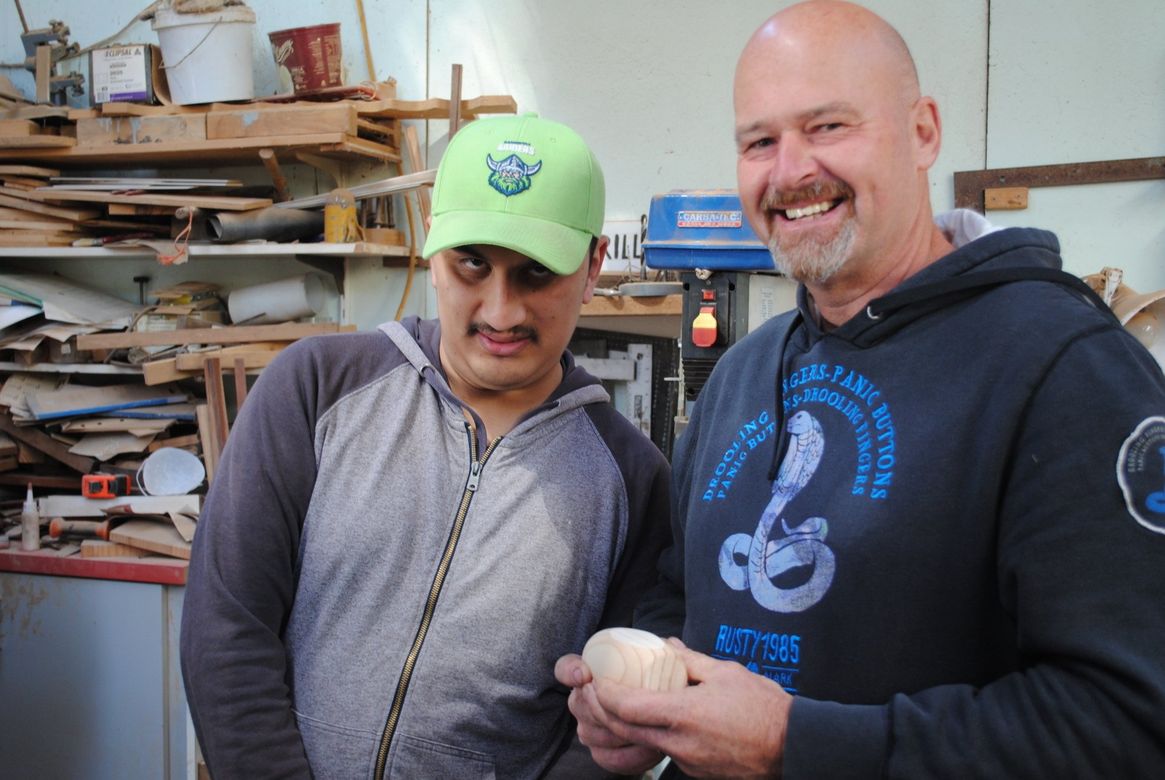 Two men smiling in a workshop, one holding a small white object, with shelves of materials behind them