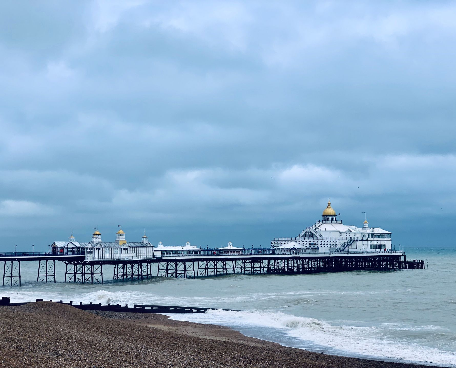 Seaside pier with a domed building under cloudy skies above a pebble beach and calm waves