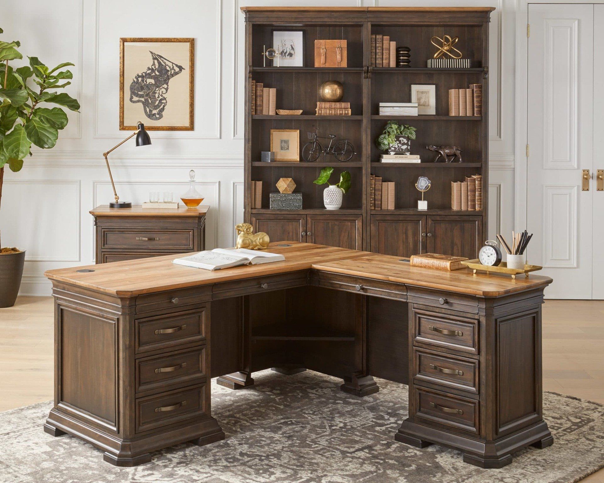 L-shaped wooden desk and bookshelf in a home office. A lamp and decorative items are also visible.