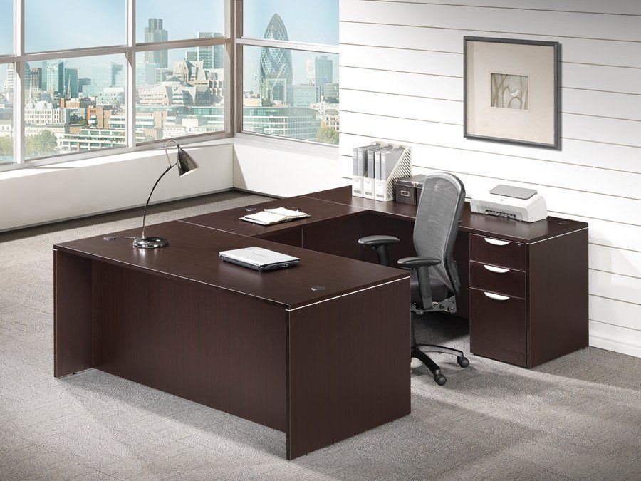 Brown U-shaped office desk with chair, lamp, and printer next to a window overlooking a cityscape.