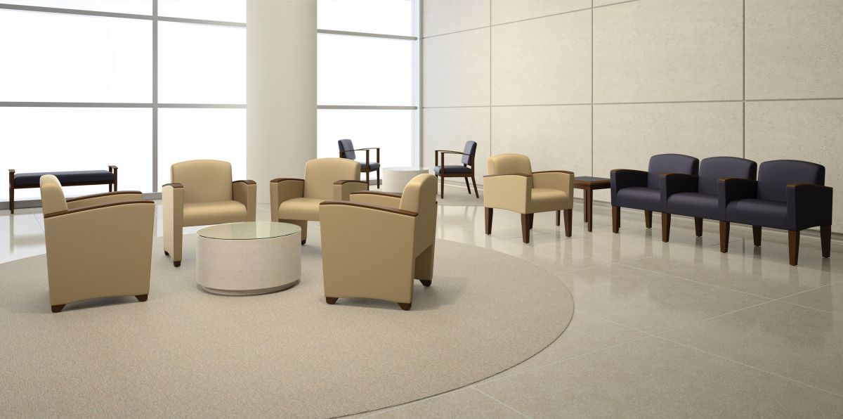 Waiting room with armchairs, couches, and round table. Beige and blue seating, large windows, and beige flooring.