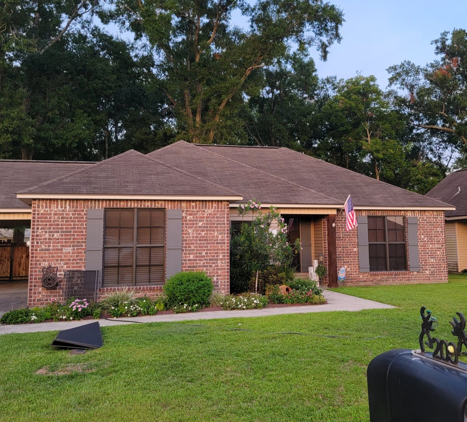 Brick house with brown roof, green lawn, trees in background, and an American flag.