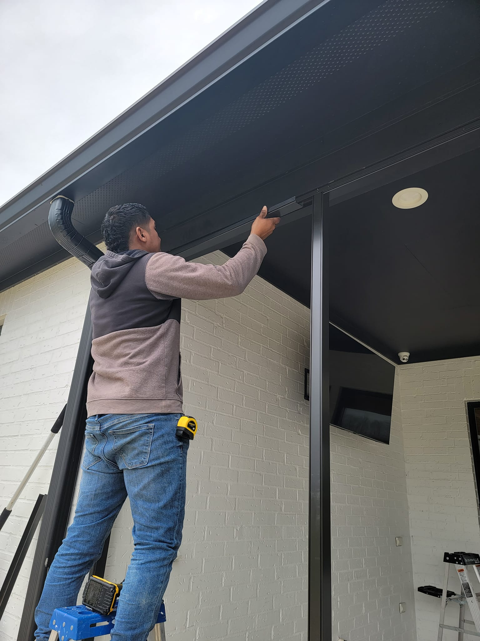 Person on a ladder installing black material on a home's exterior, white brick wall.