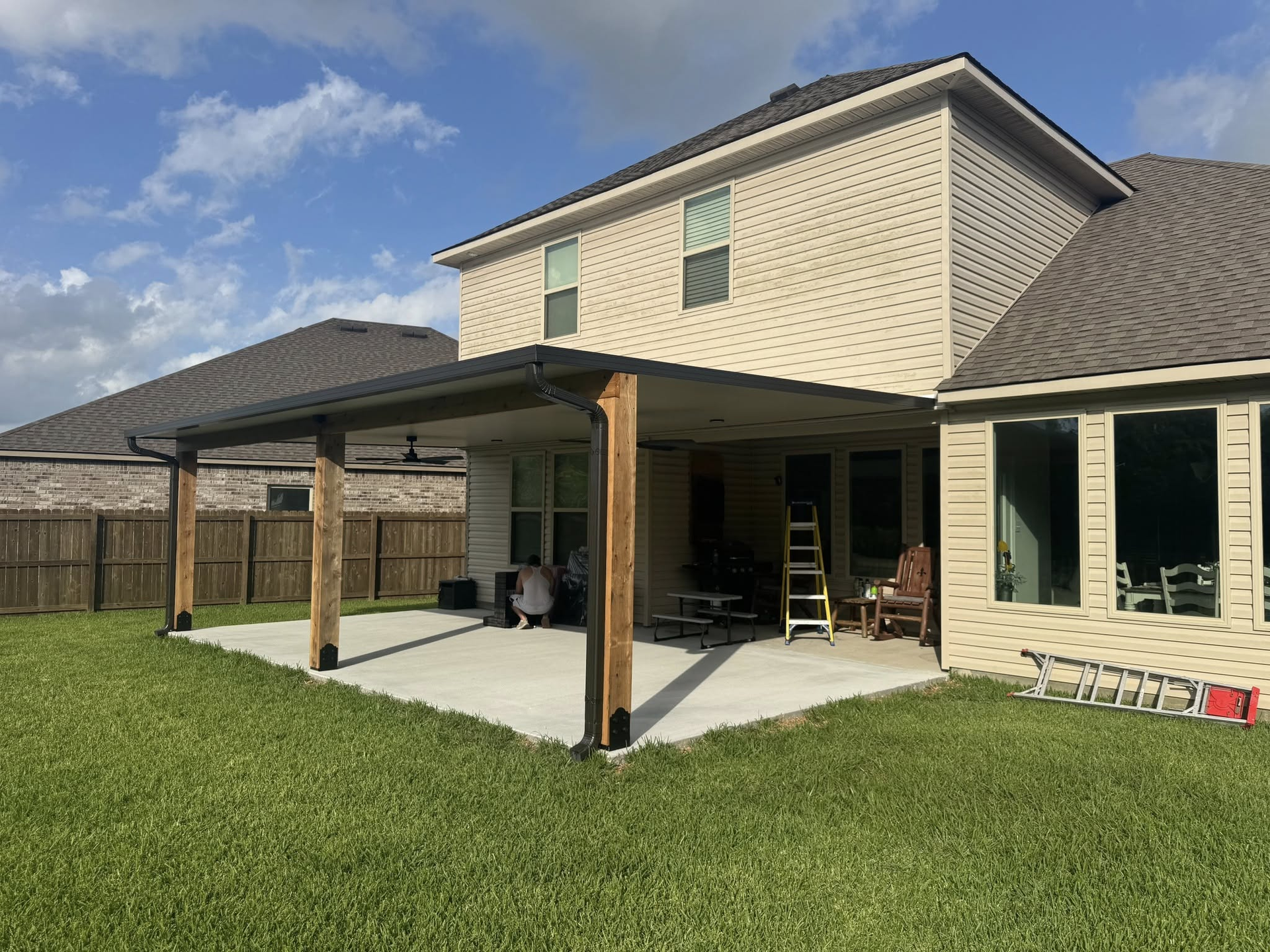 Backyard patio with a concrete slab, wooden posts, and a dark roof extension. Green lawn and brick house in the background.