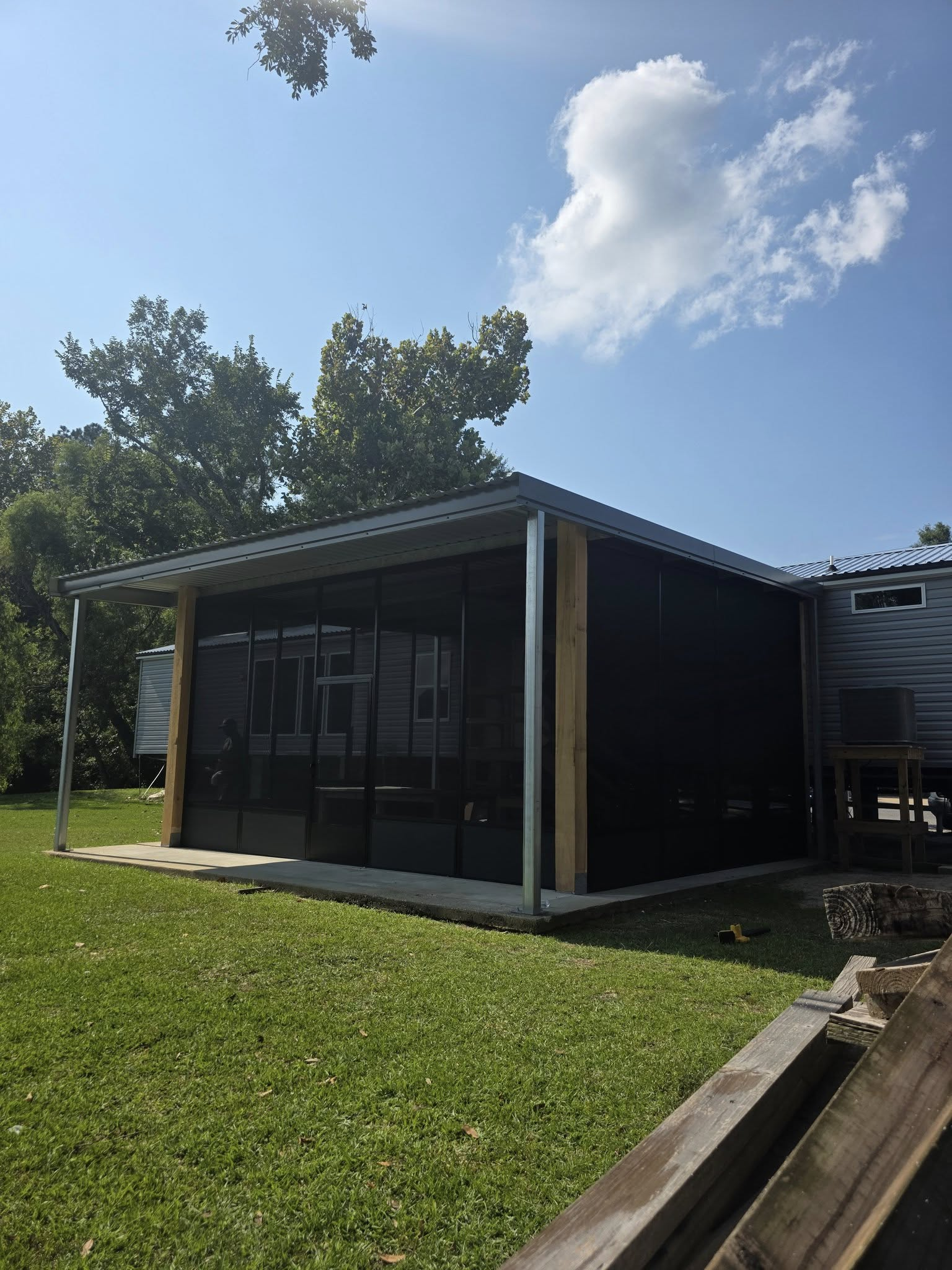 Screened-in porch with black screens, metal roof, and wooden posts on a grassy lawn under a blue sky.