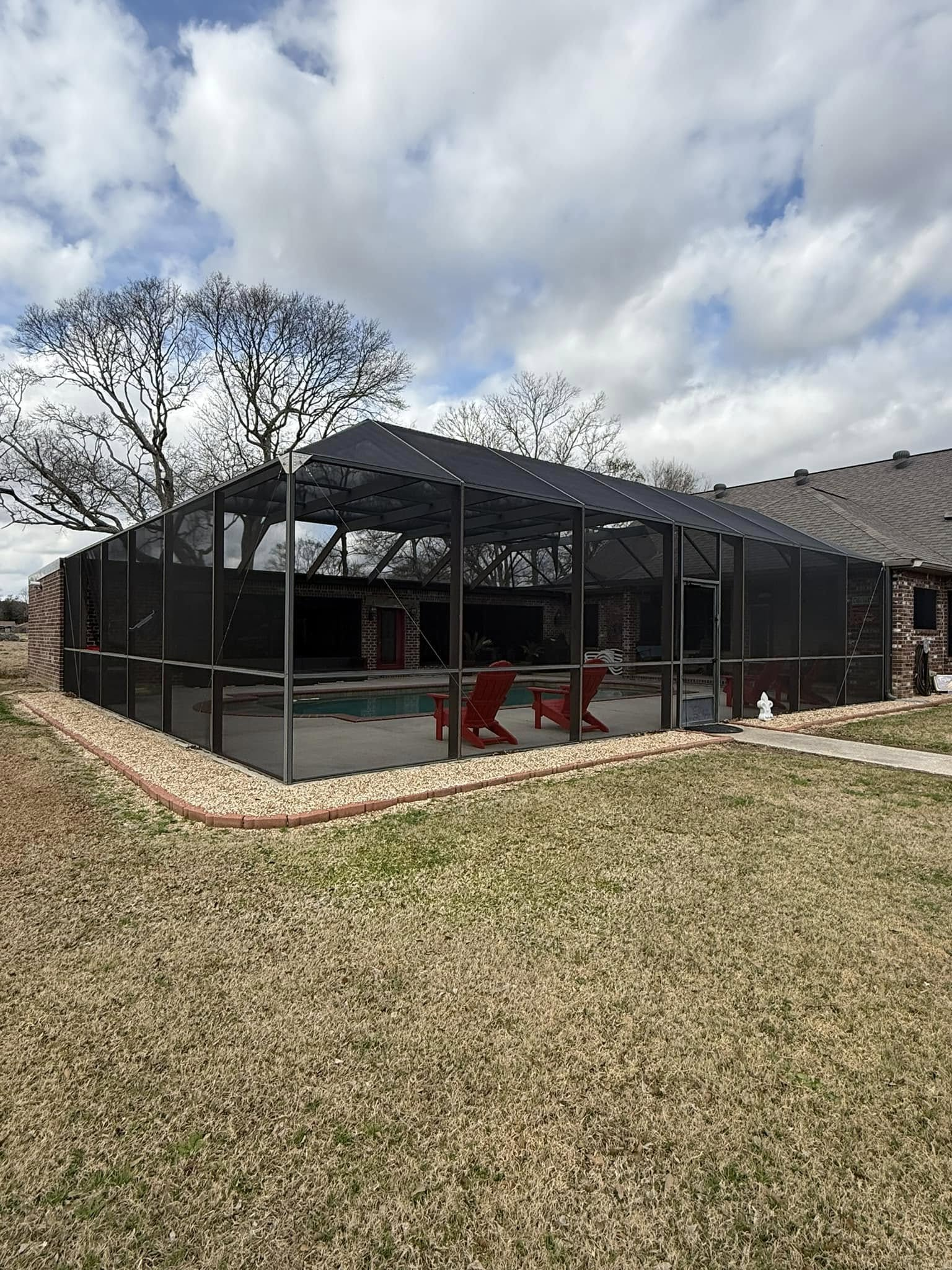 Screened pool enclosure with red chairs, on a grassy lawn under a cloudy sky.