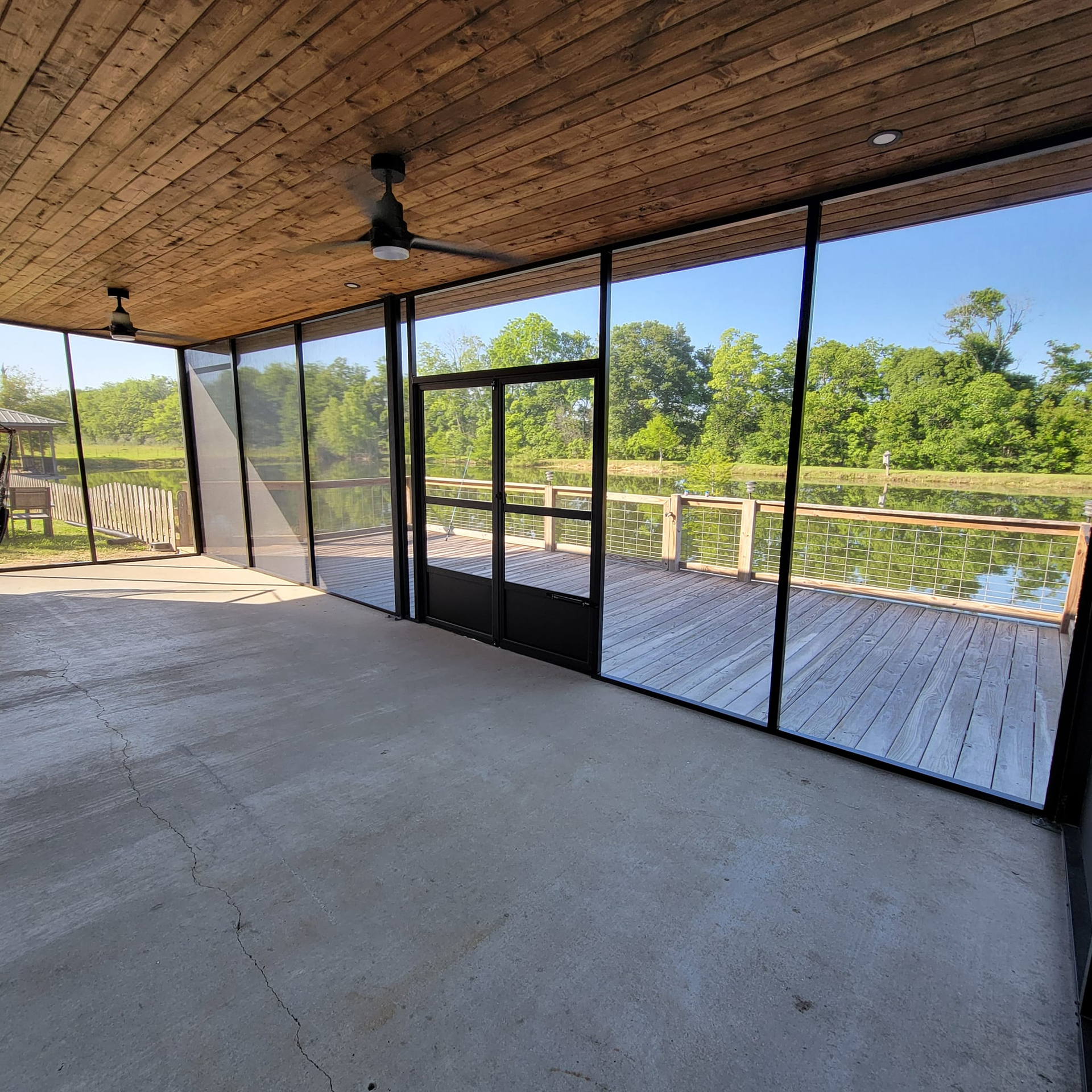 Screened-in porch overlooking a lake, with wooden ceiling, concrete floor, and black-framed screens and door.