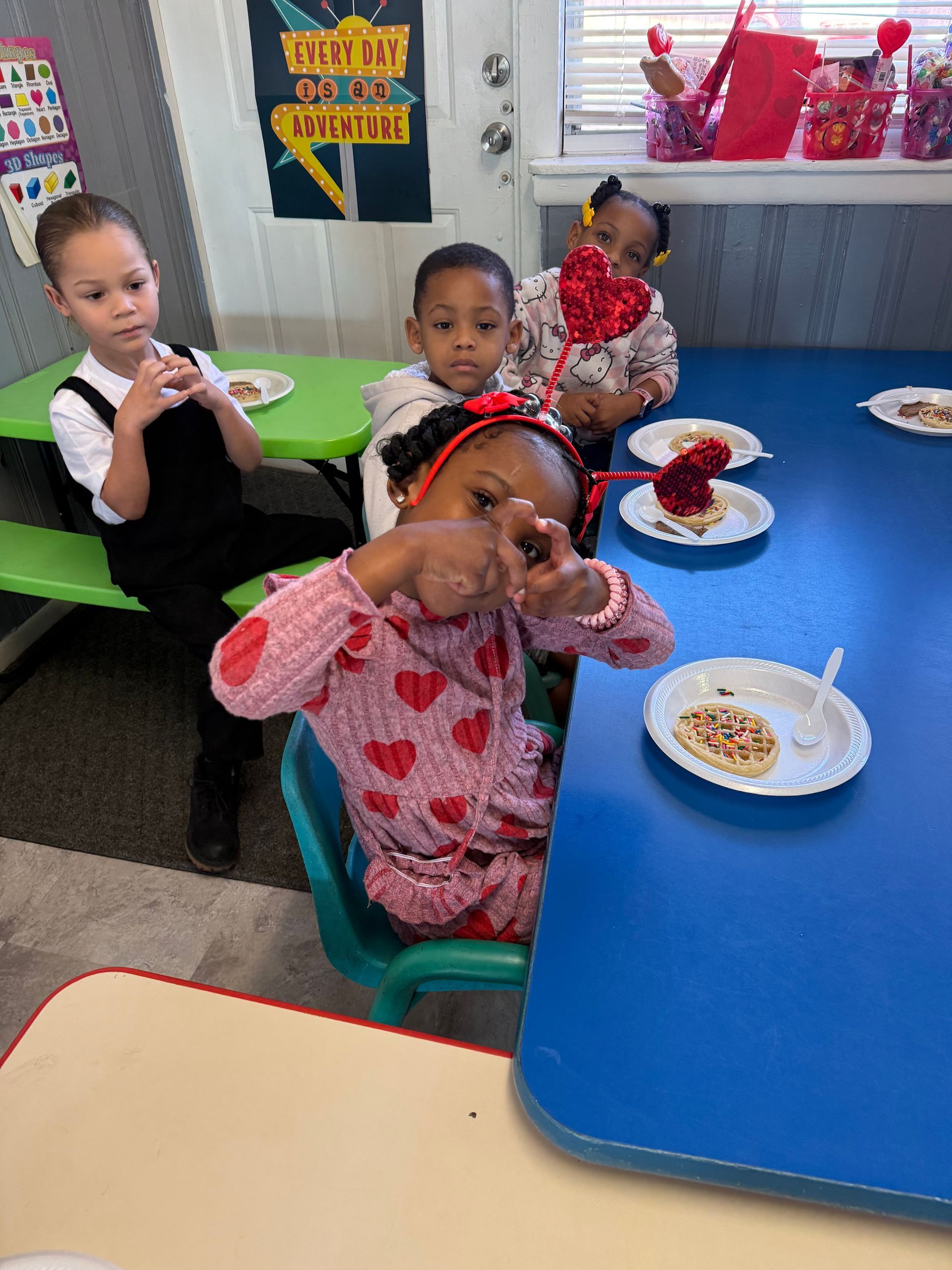 Children at a table with Valentine's treats; one girl makes a heart with her hands.