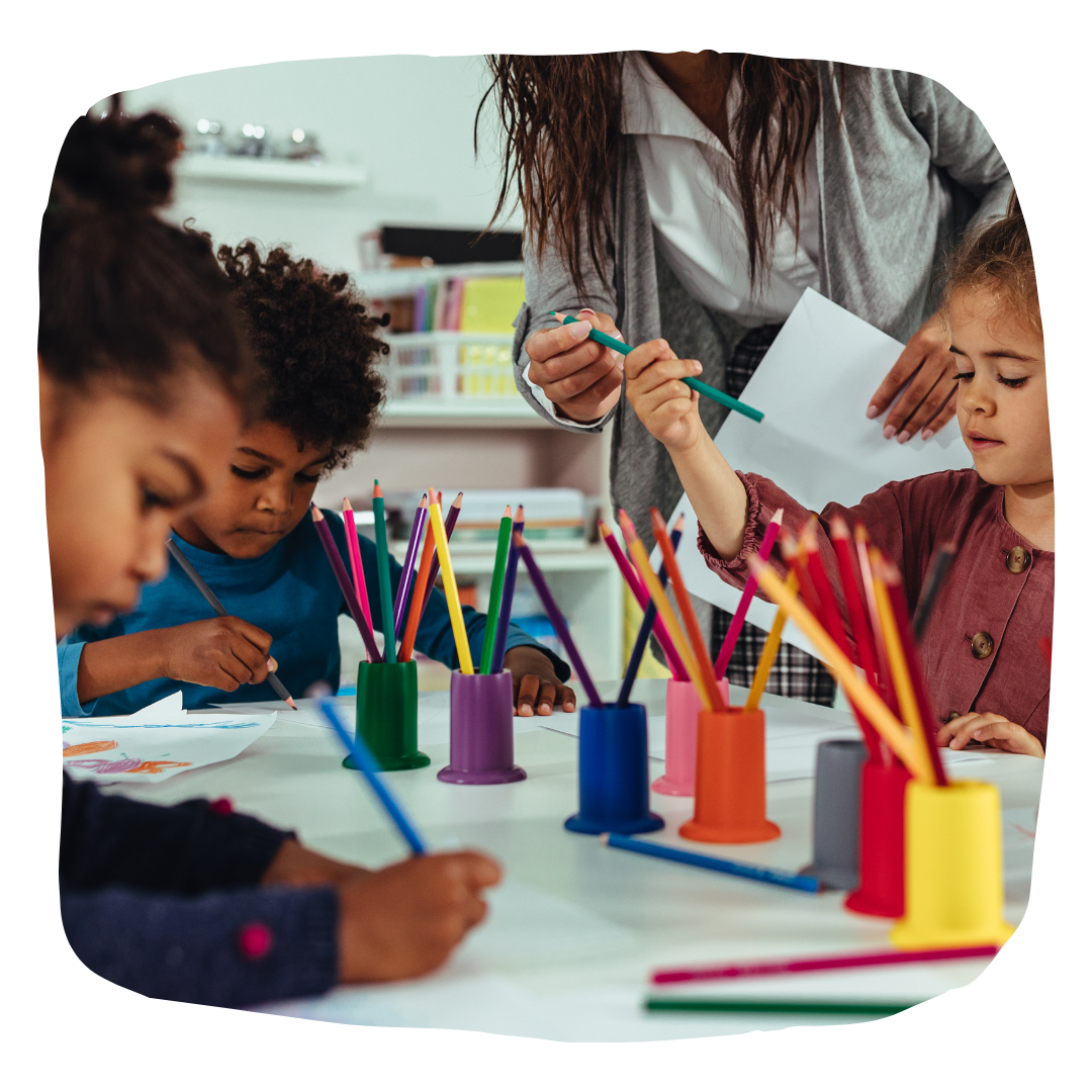 Children drawing at a table with a teacher holding paper. Crayons in colorful cups.