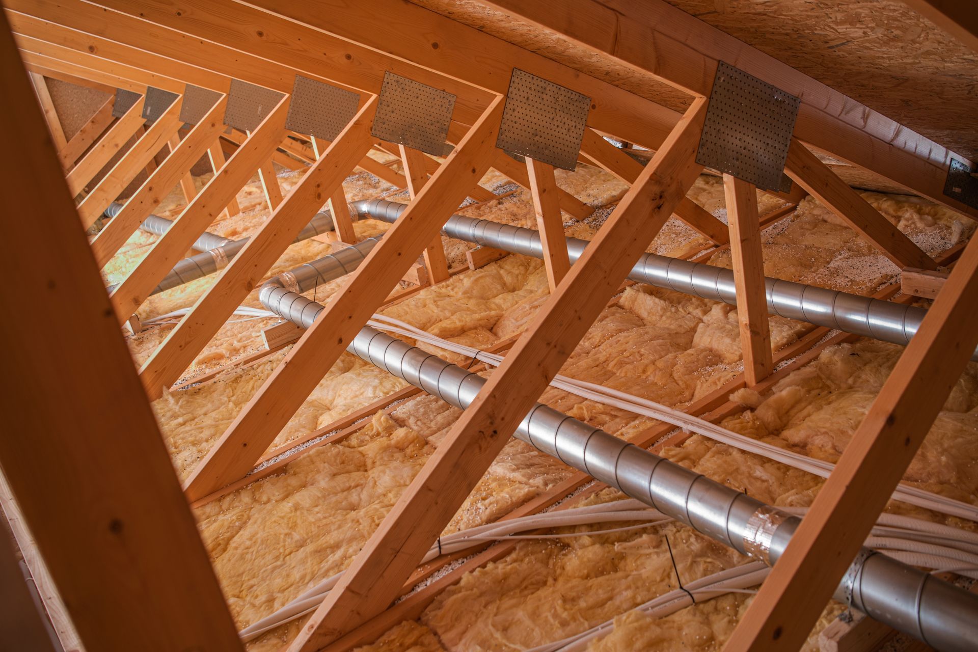 Wooden roof trusses and metal ductwork inside an unfinished attic with insulation