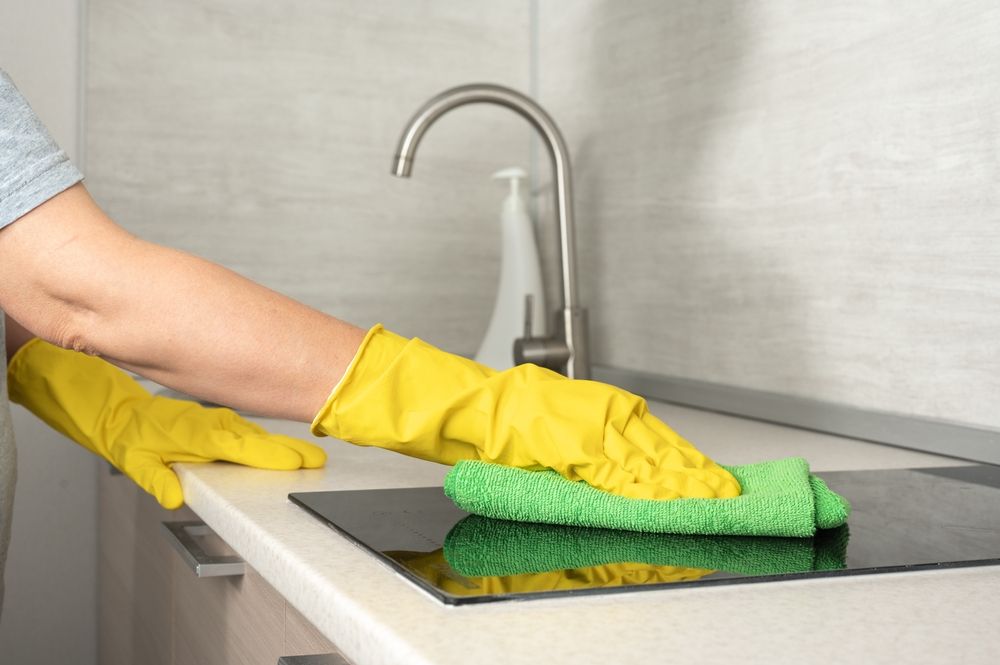 A person wearing yellow rubber gloves cleans a black induction stovetop with a green cloth in a modern kitchen.