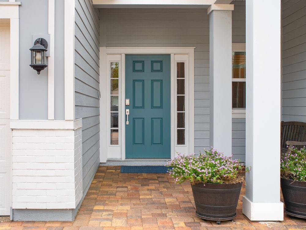 A teal front door with side windows, flanked by gray siding, a wall-mounted light, and potted plants on a brick porch.