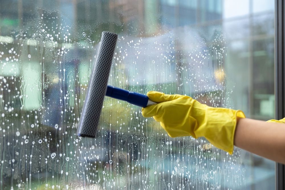 A hand in a yellow rubber glove holds a squeegee to clean soap suds off a glass window.