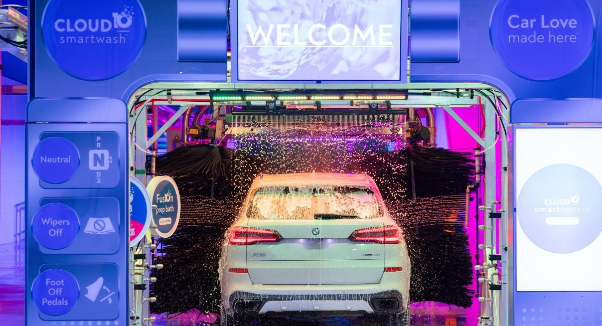 Car wash bay with a white SUV under a colorful “WELCOME” sign and spraying water