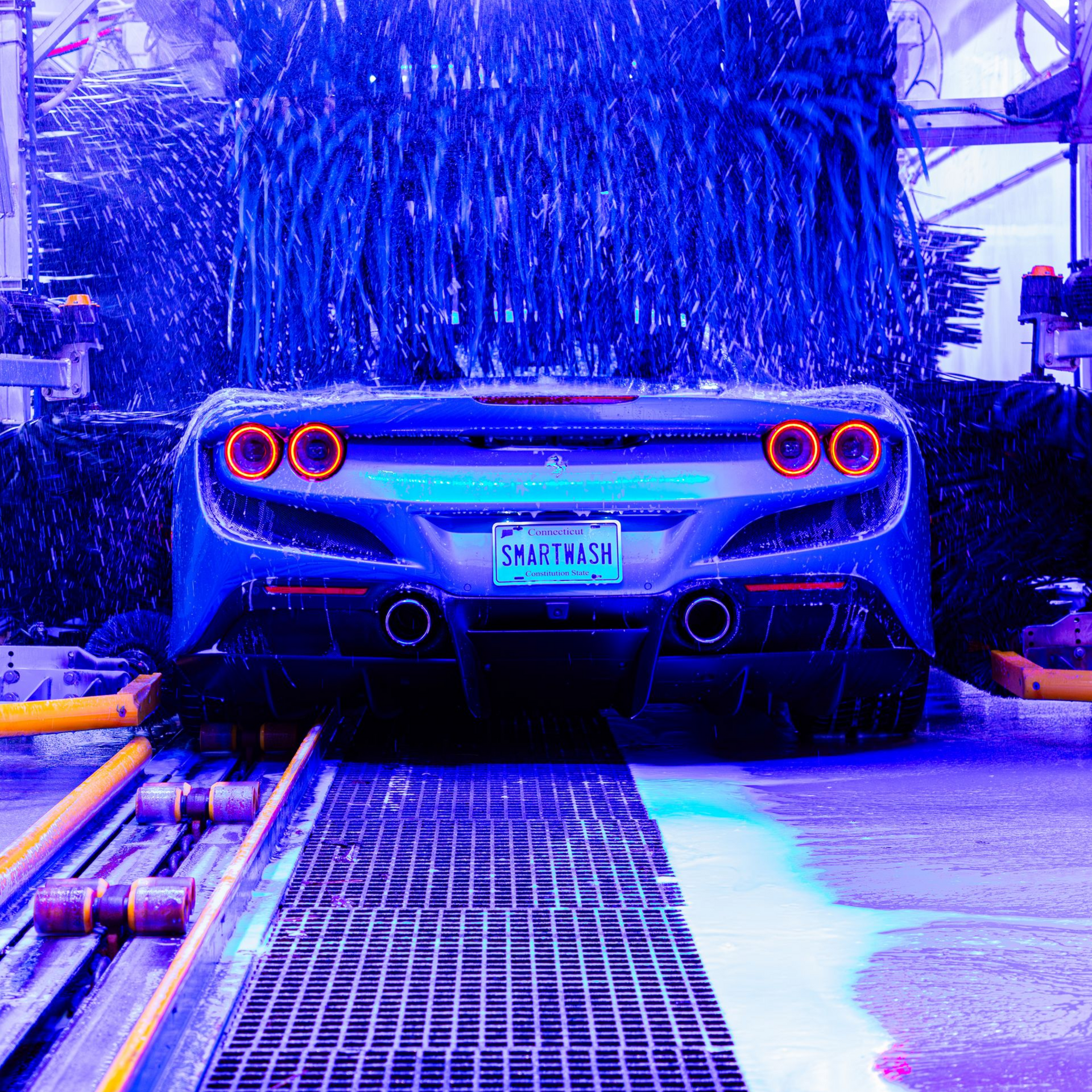 Rear view of a silver sports car with glowing taillights, parked in a wet car wash under blue lighting.