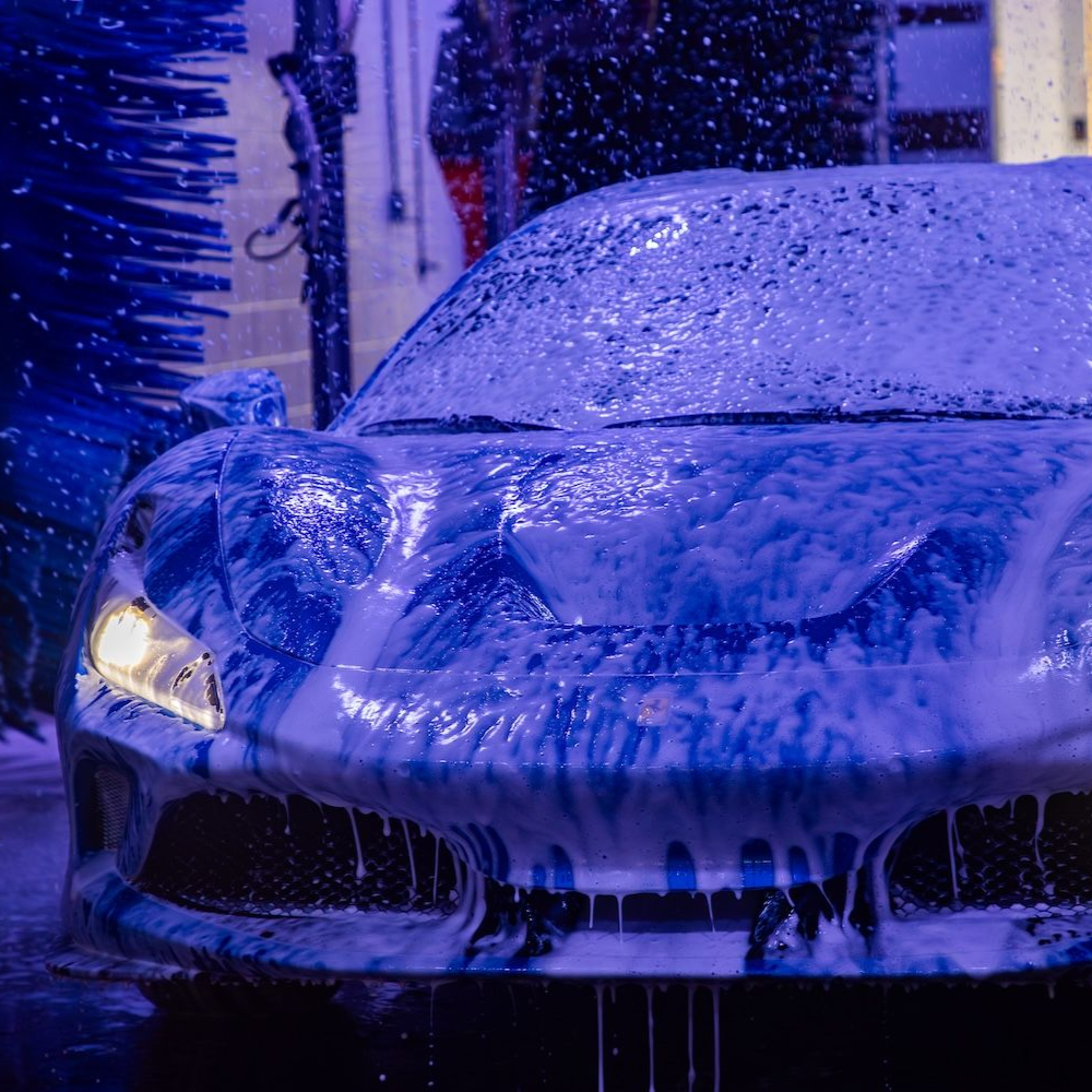 Snow-covered car in a blue-lit car wash, front headlights glowing.
