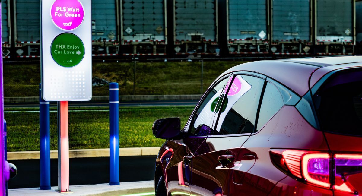 Dark sedan at a parking or charging station with a sign and blue posts in front of a building.