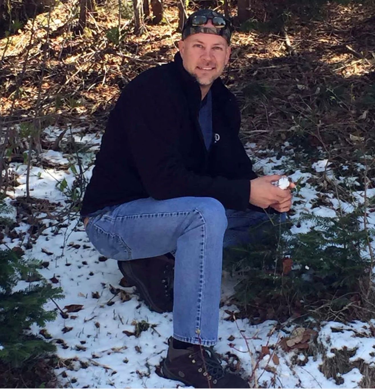 Man crouching in snowy woods, wearing a black jacket, jeans, and a cap.