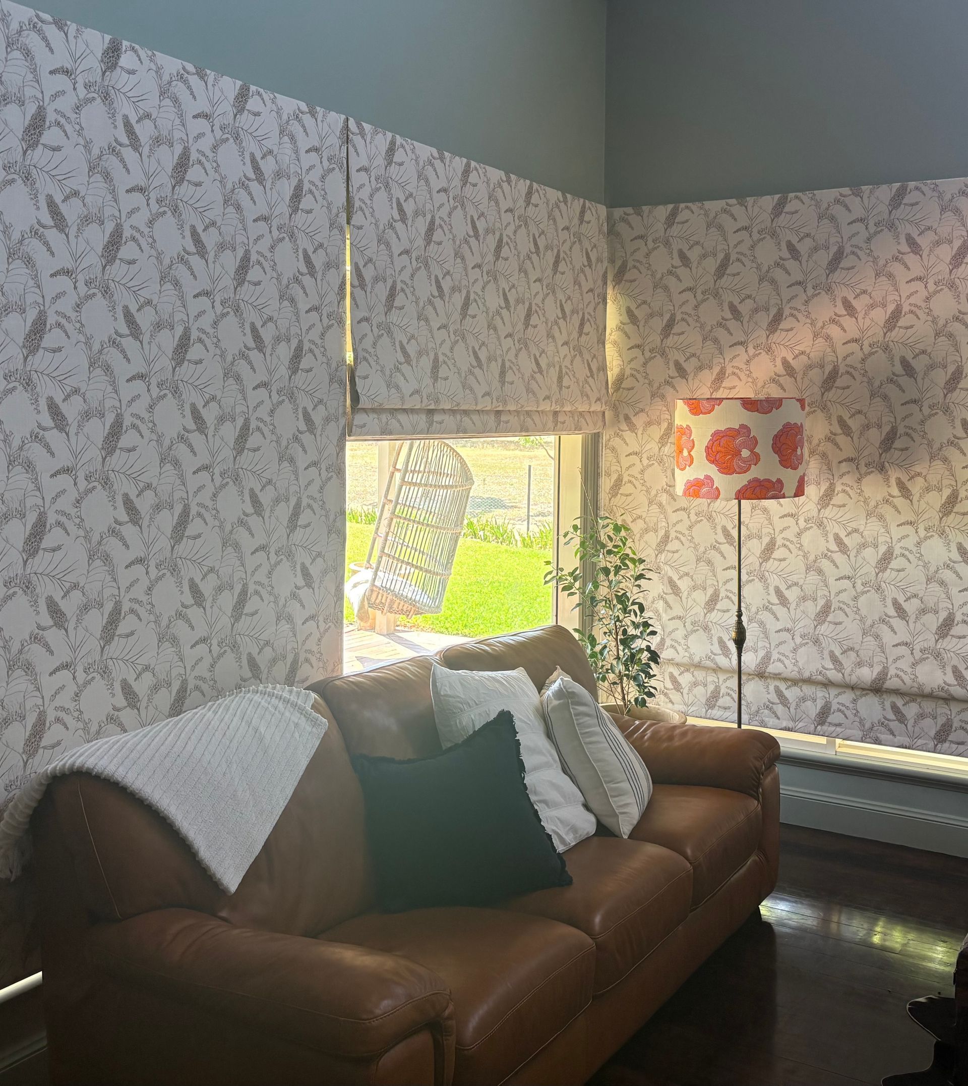 A brown leather sofa sits in a room with patterned wallpaper, window shades, and a lamp with a patterned shade.