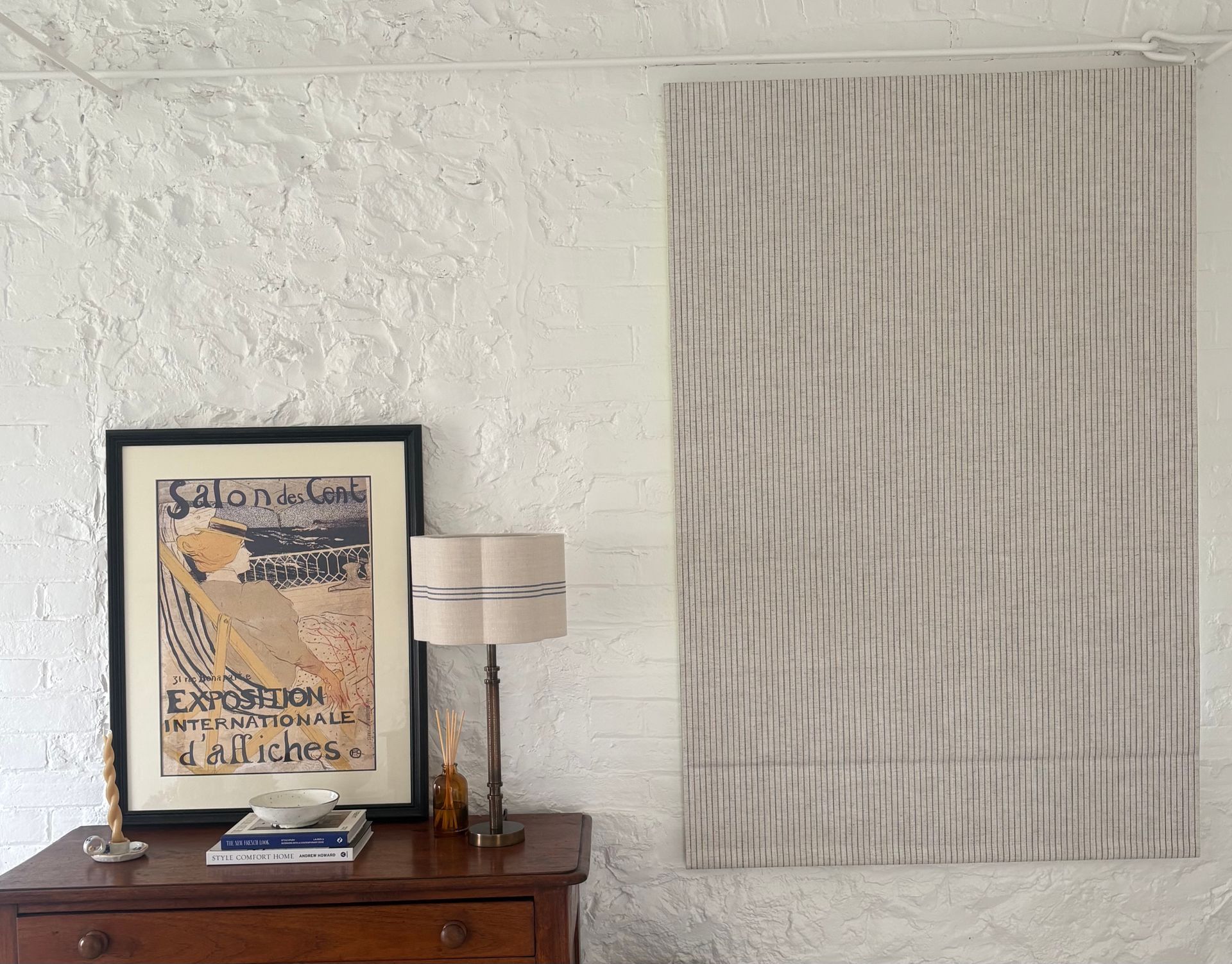 A white brick wall with a framed poster and a lamp on a wooden dresser, next to a vertical striped fabric wall hanging.