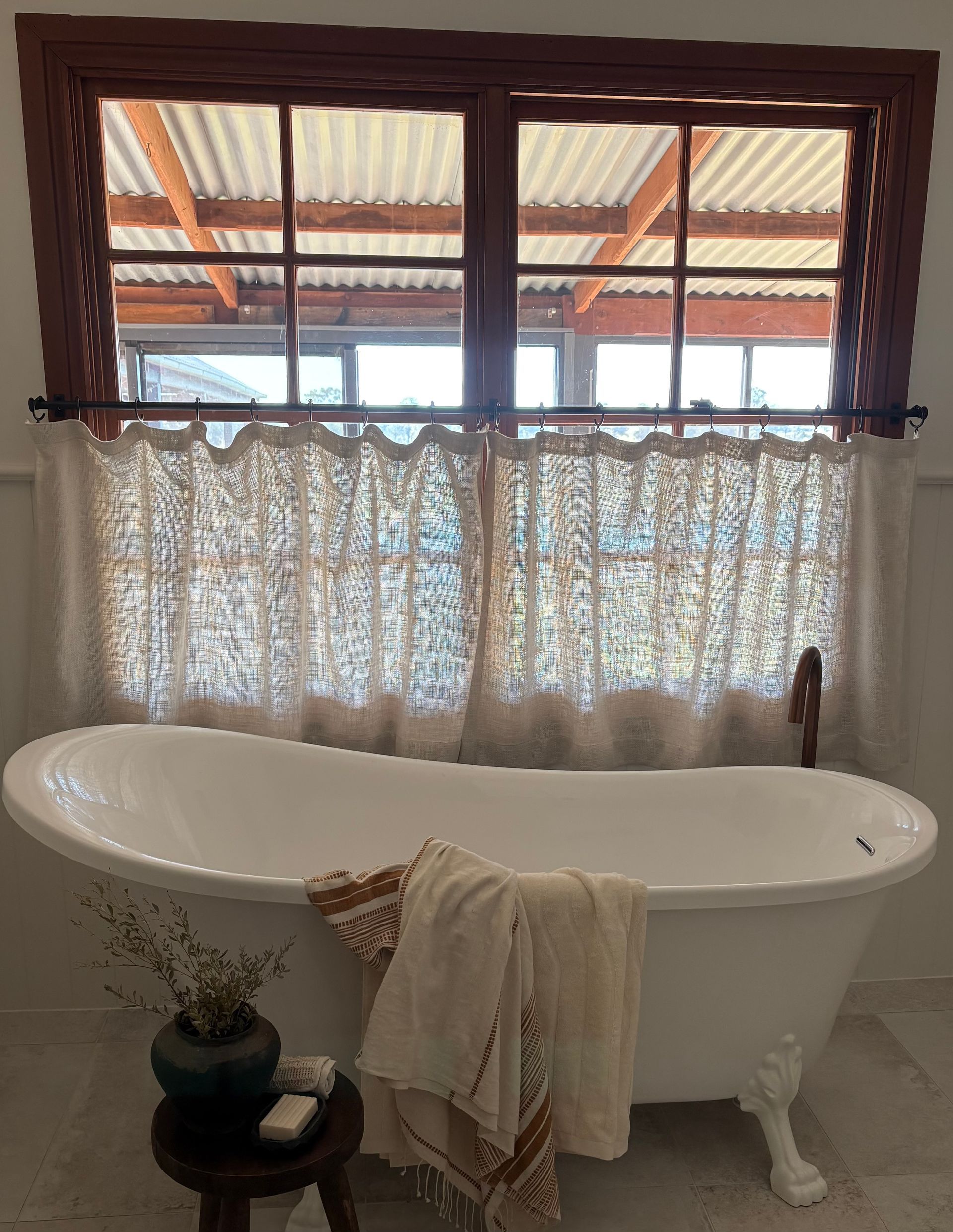 A white clawfoot bathtub sits beneath a window with lace cafe curtains, next to a small wooden stool with a vase and towel.