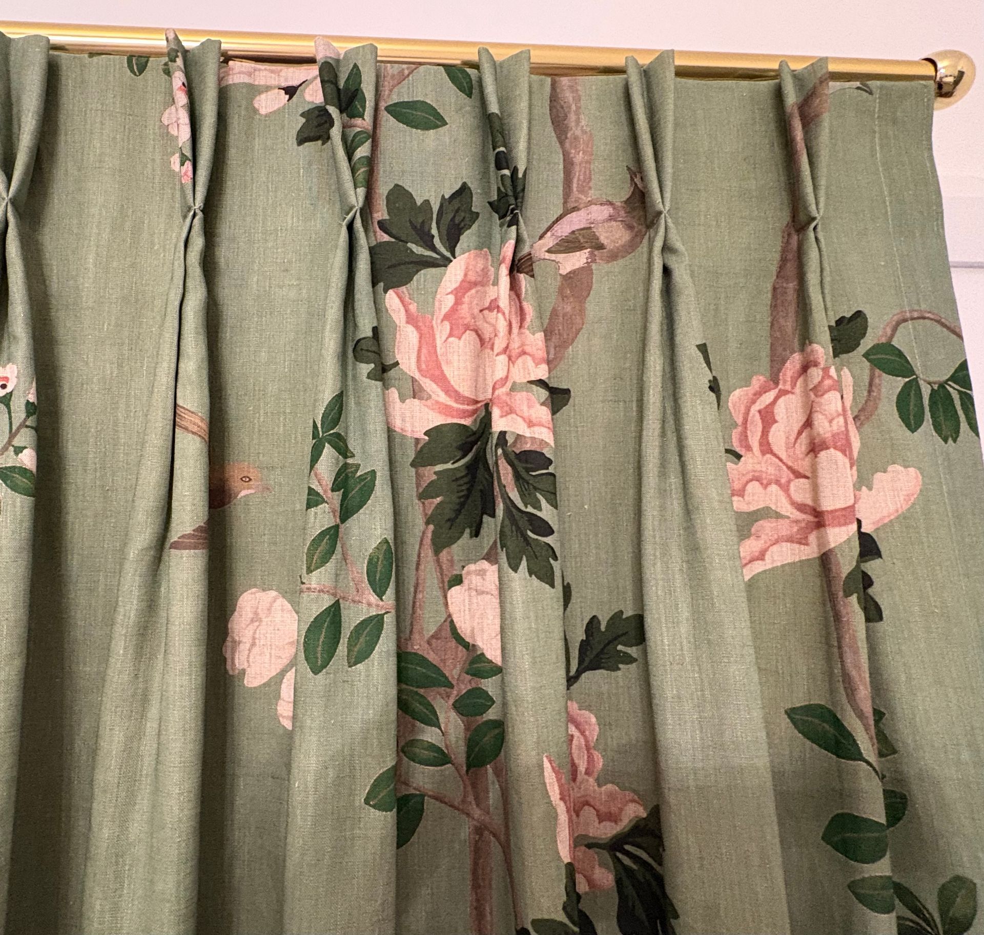 Sage green curtains with a pink floral and green leaf pattern, hung with pinch pleats on a brass curtain rod.