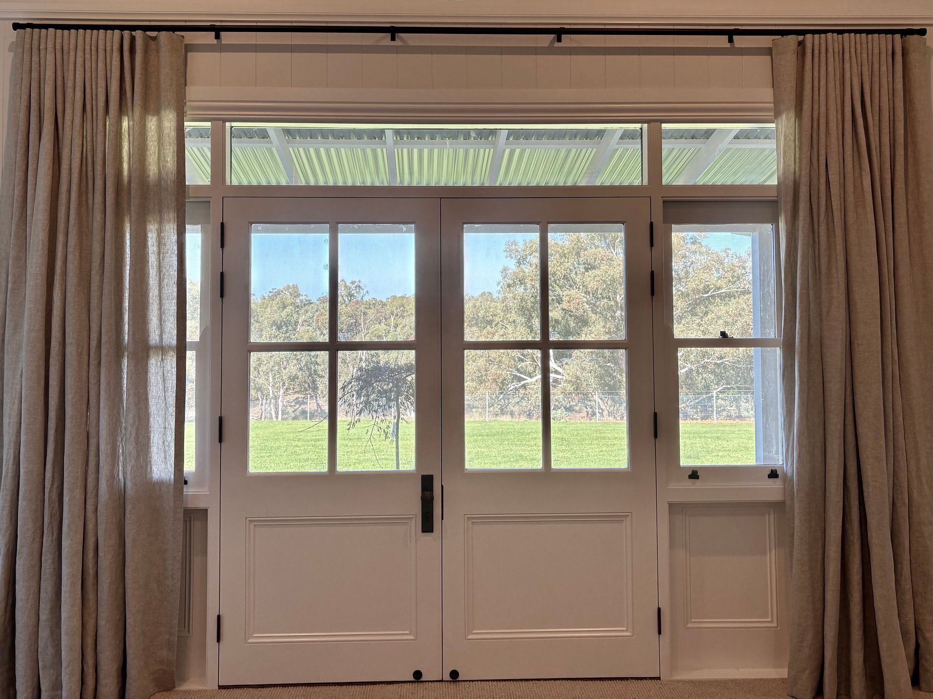Double doors with glass panes and side windows, framed by full-length beige curtains on a dark rod against a garden view.