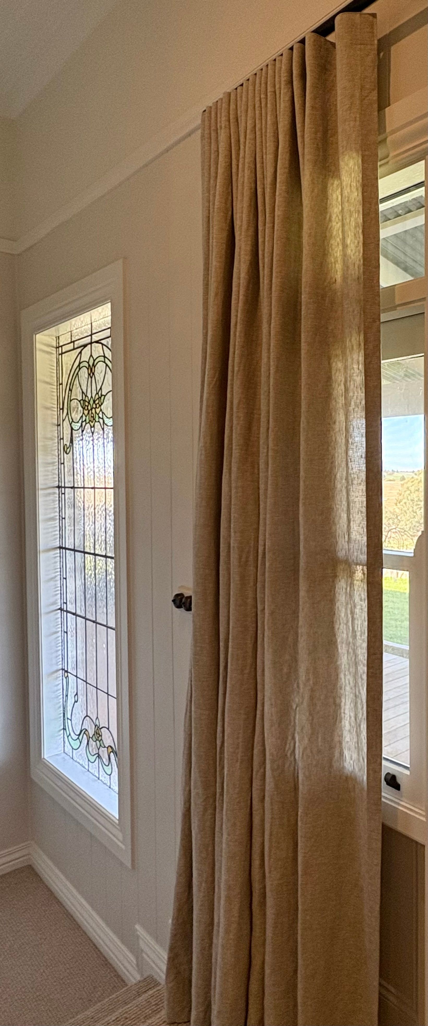 A tall, tan linen curtain hangs on a rod beside a rectangular window with a decorative metal grate.