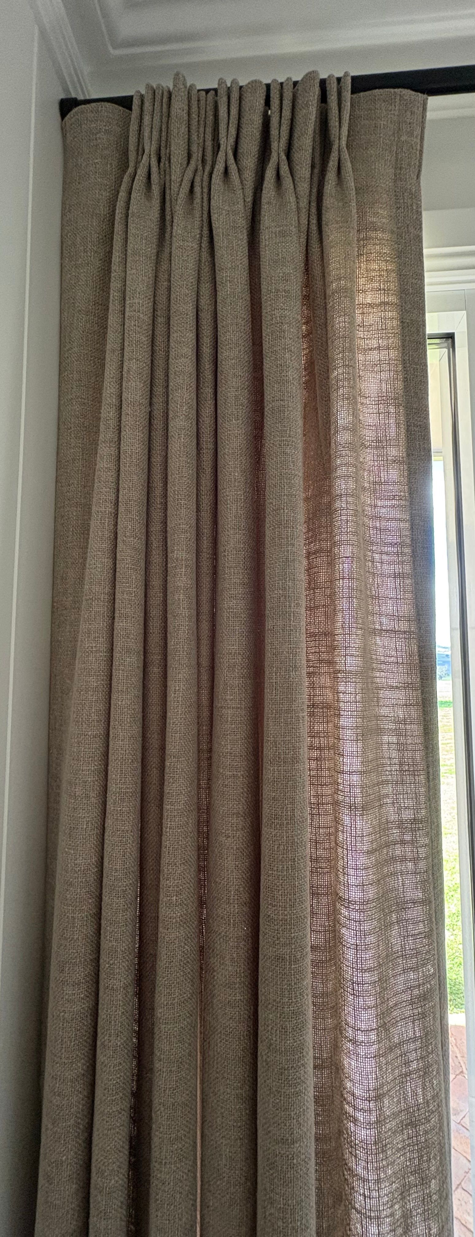 Close-up of a pleated beige curtain hanging on a black rod against a light wall, with a sheer panel visible on the right.