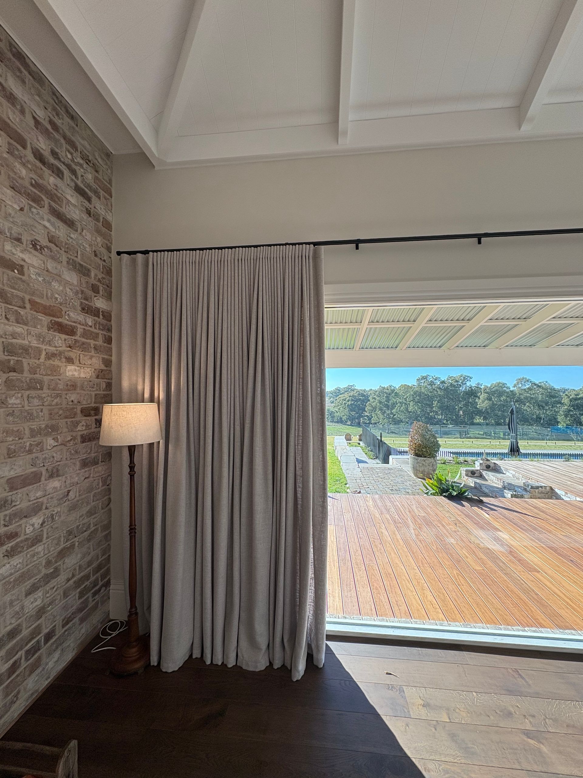 A tall floor lamp stands next to light-colored floor-to-ceiling curtains in a room with a rustic brick wall and patio view.
