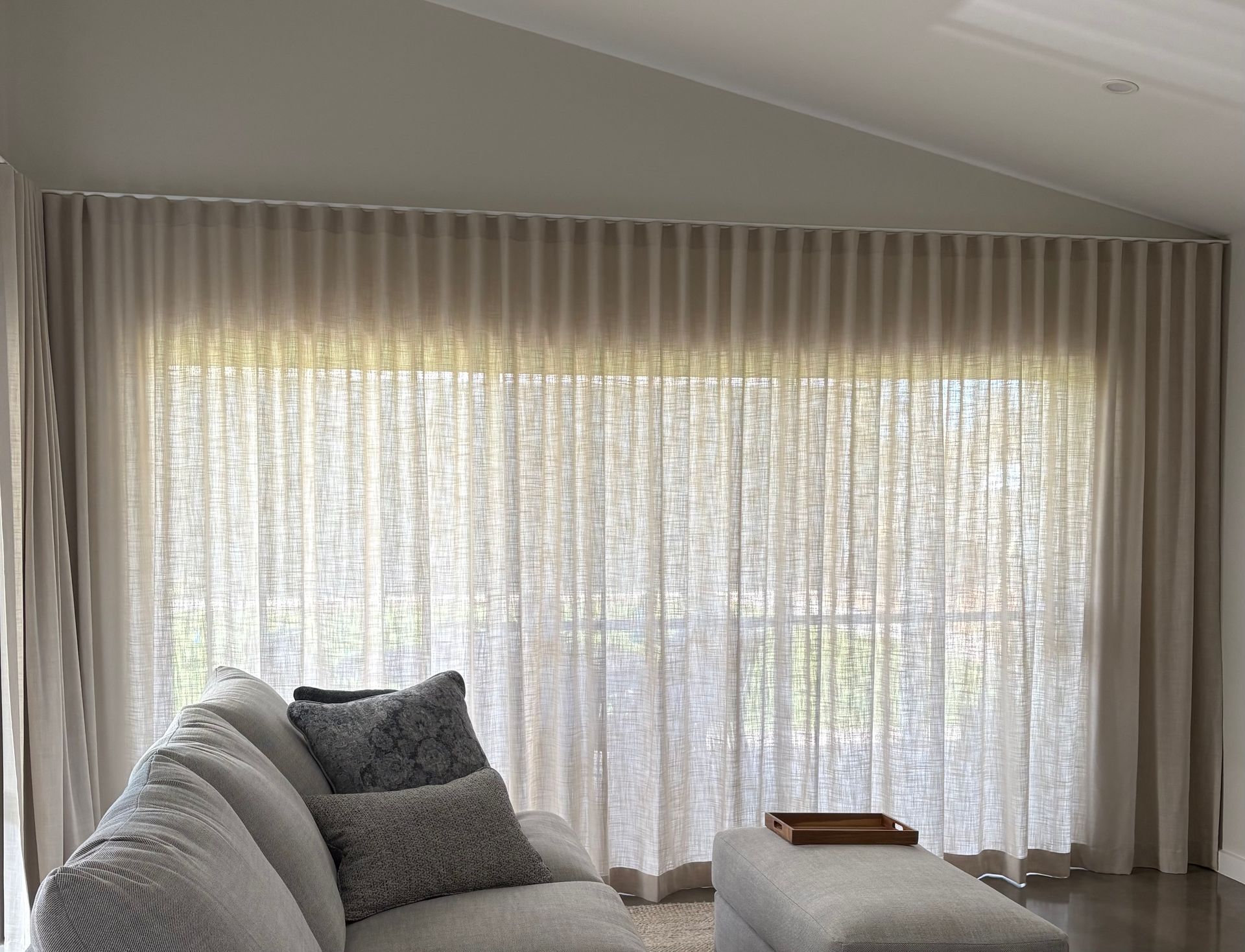 Floor-to-ceiling beige curtains with a pleated wave fold hang over a large window next to a gray sofa and ottoman.