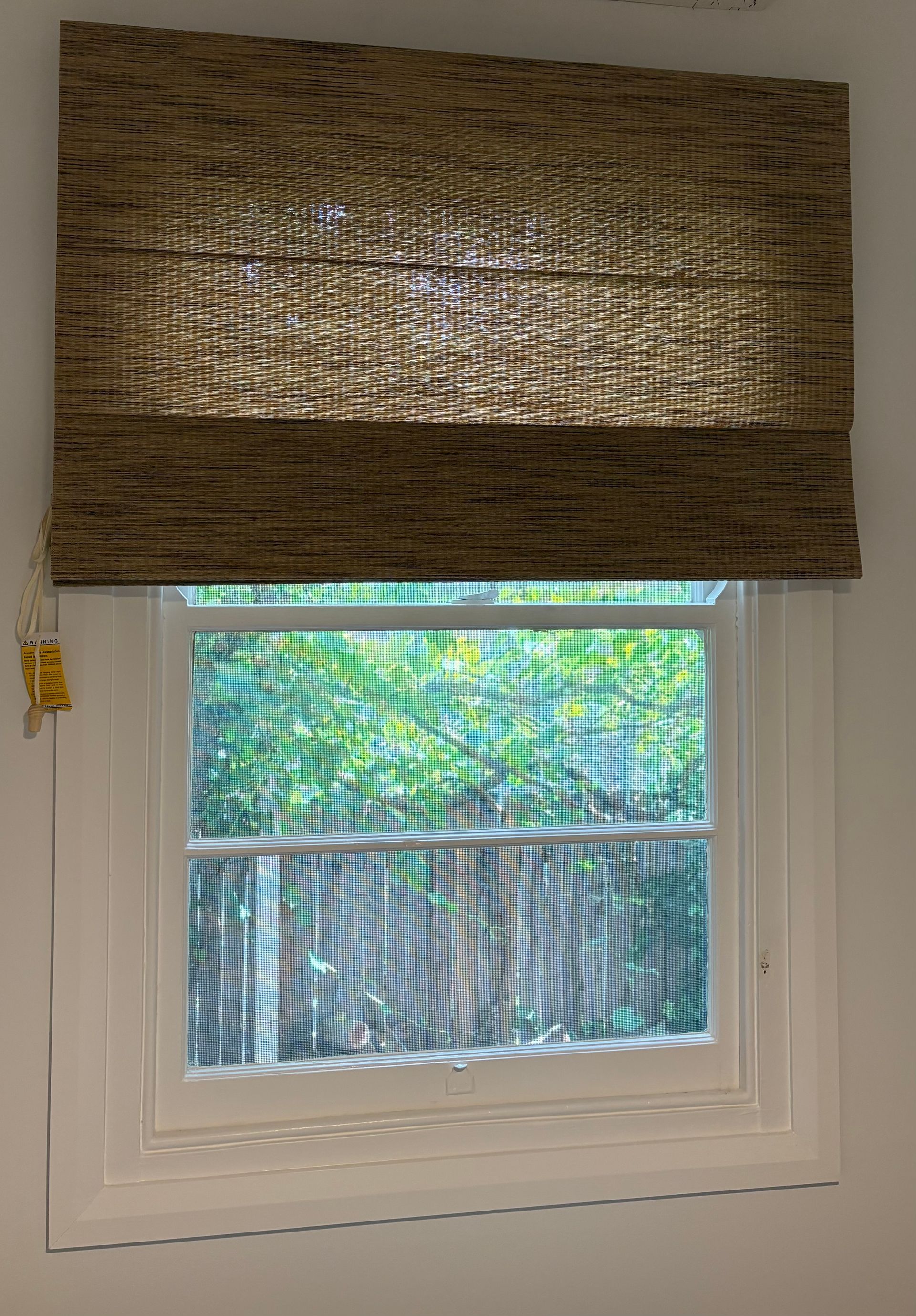 A woven bamboo Roman shade partially covers a white-framed window looking out onto a green backyard.