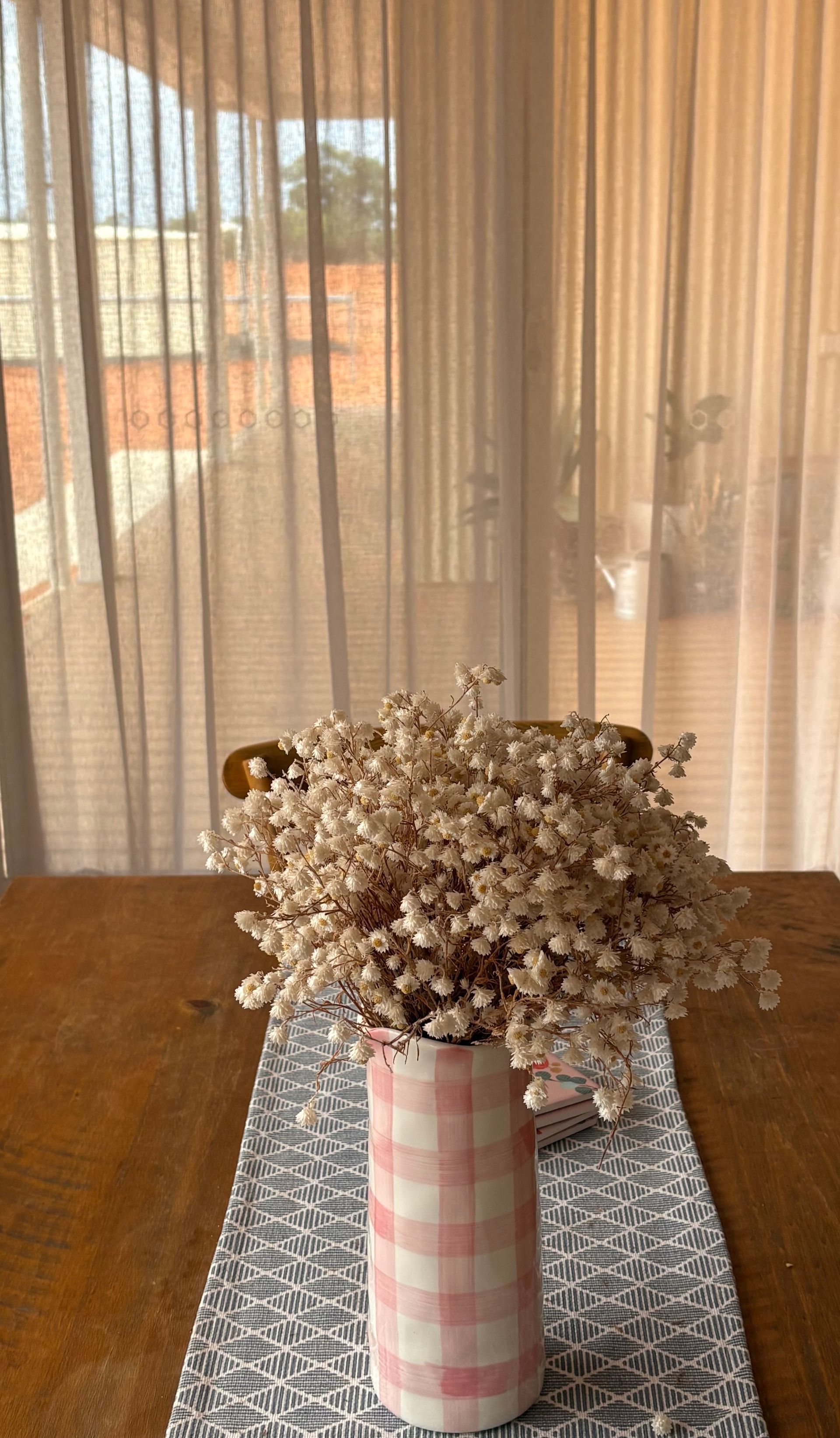 A vase with a pink-and-white gingham pattern filled with dried white flowers, sitting on a table with a patterned runner.
