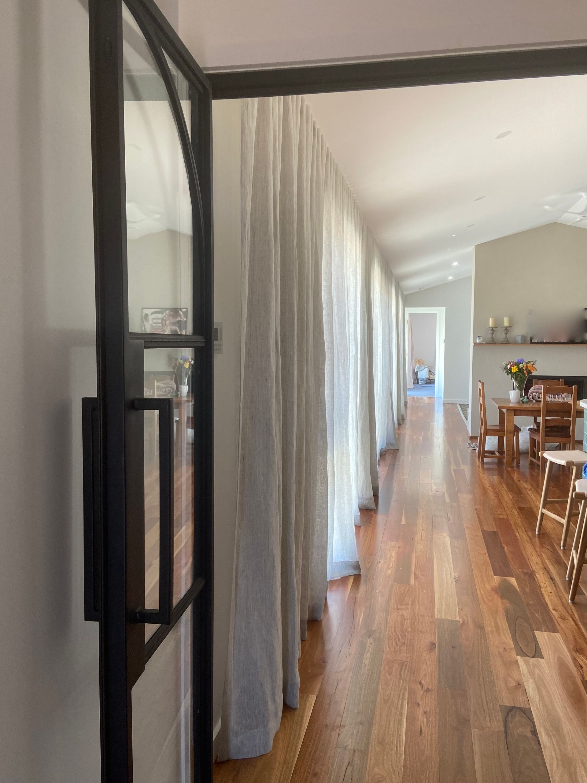 A partial view of a black-framed glass door opening into a hallway with sheer curtains, wood floors, and a dining area.