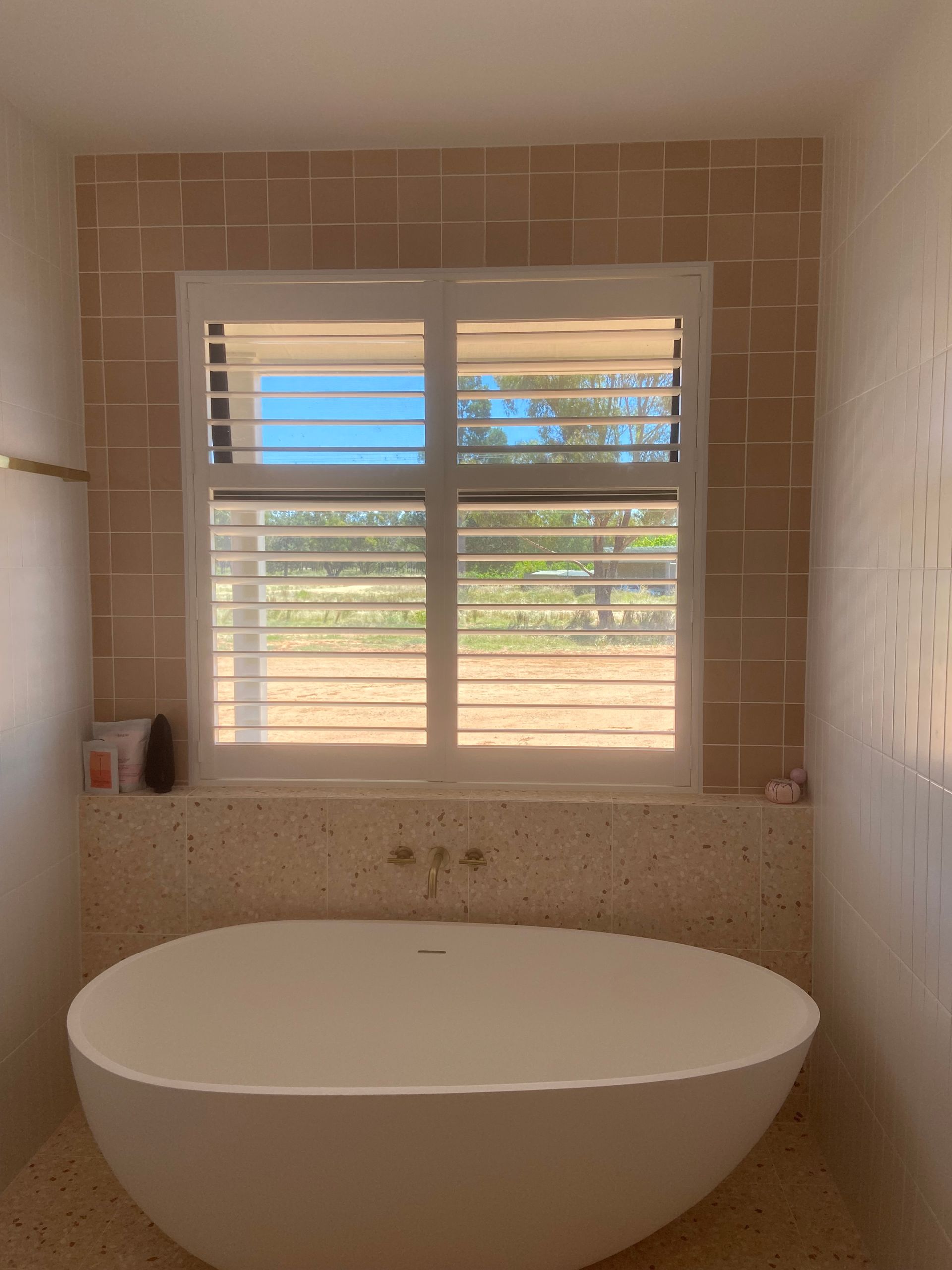 A white, oval freestanding bathtub sits in front of a window with white shutters, surrounded by tan tile walls and floors.