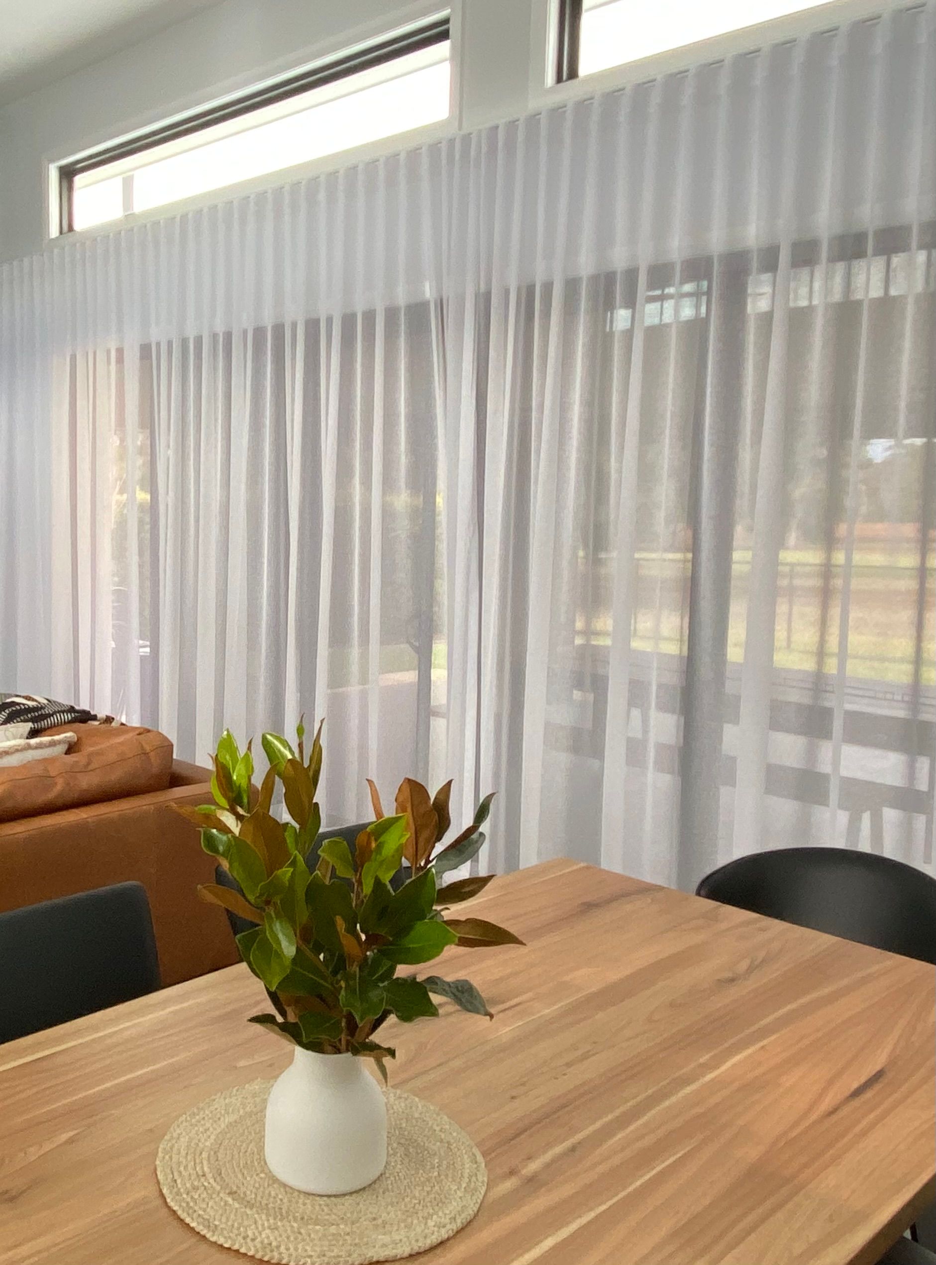 A wooden dining table with a potted plant sits in front of a wall of tall, white, floor-to-ceiling sheer curtains.