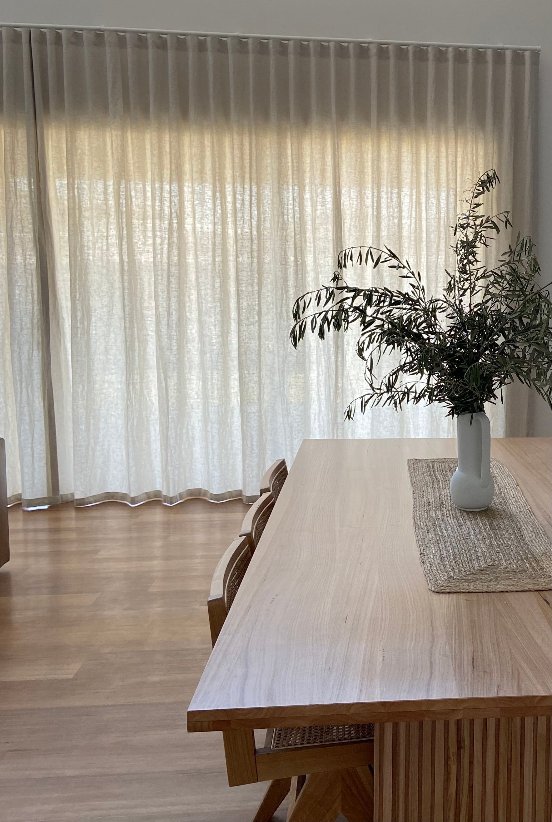 A light wood dining table with a vase of greenery and a runner sits near floor-to-ceiling sheer curtains in a bright room.
